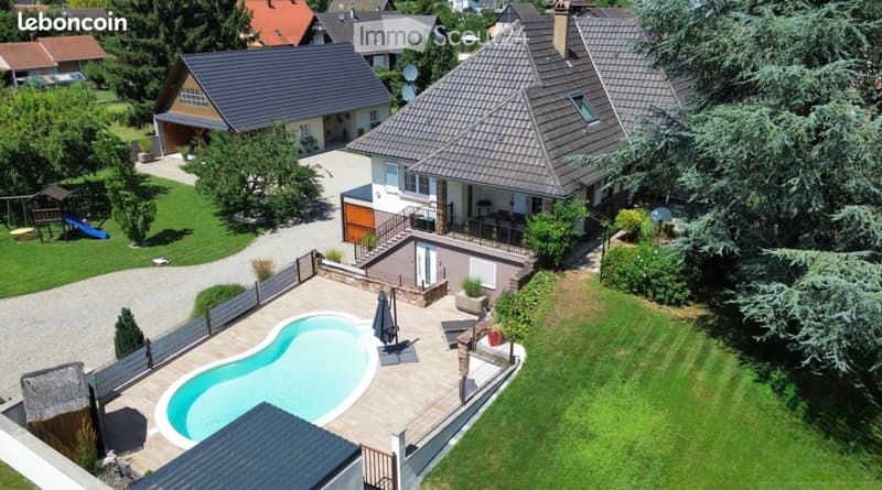 Aerial view of a two-story house with a tiled roof, surrounded by a well-maintained garden, featuring a swimming pool, patio, and outdoor furniture.