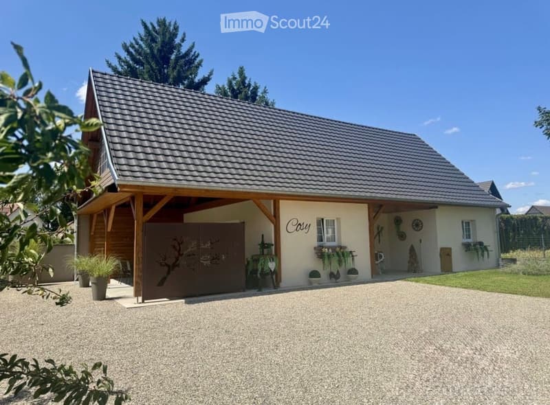 Single-story house with a tiled roof, covered entrance area, and a gravel driveway. The exterior features light-colored walls, decorative elements, and potted plants.