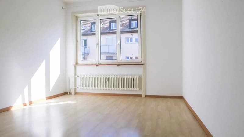 Empty room with light wooden flooring, white walls, and a large window with a view of neighboring buildings. A radiator is visible below the window.
