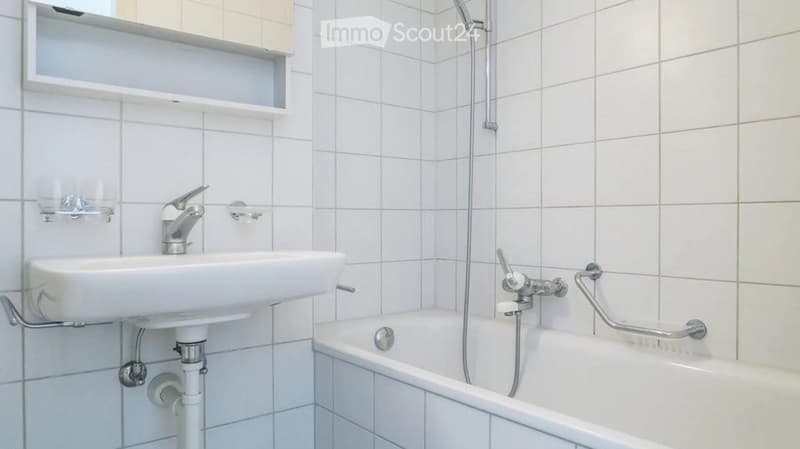 A small bathroom with white tiled walls and floor, featuring a sink with a faucet, a bathtub with a handheld showerhead, and a handrail for support.