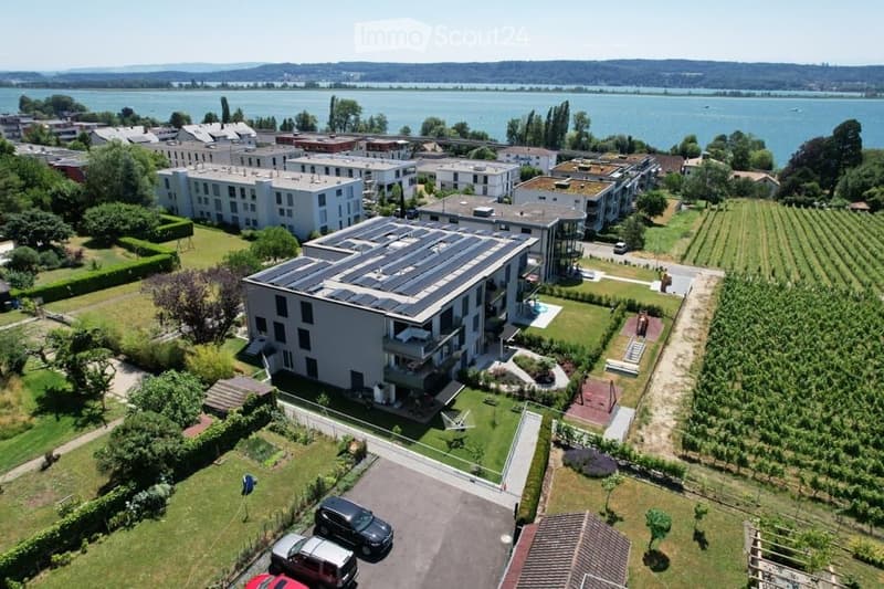 Aerial view of a modern residential complex with multiple buildings, solar panels on the roof, a swimming pool, and a scenic lake view in the background.