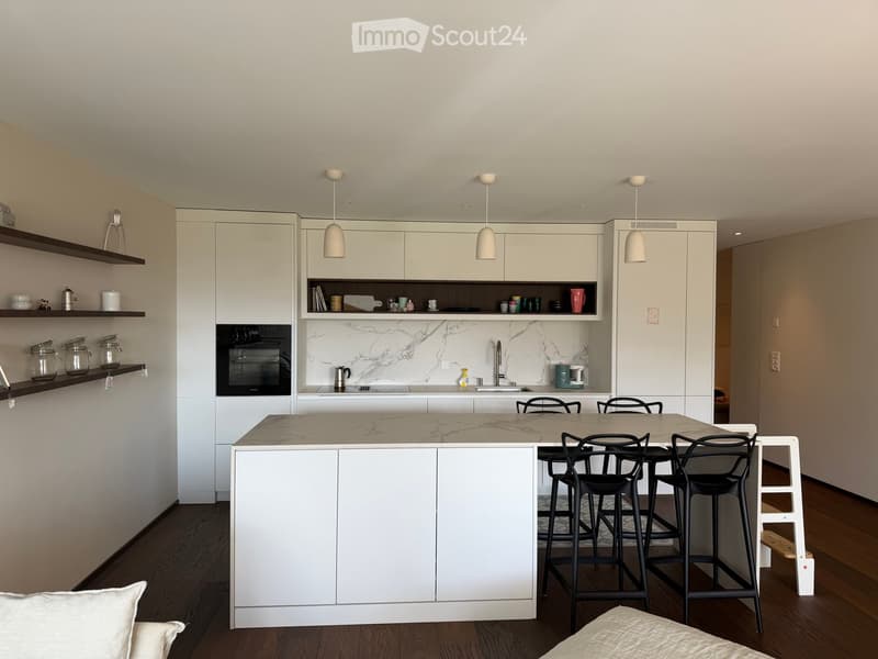 Modern kitchen with white cabinets, marble countertops, central island, bar stools, and pendant lights. Shelves hold jars and cups.