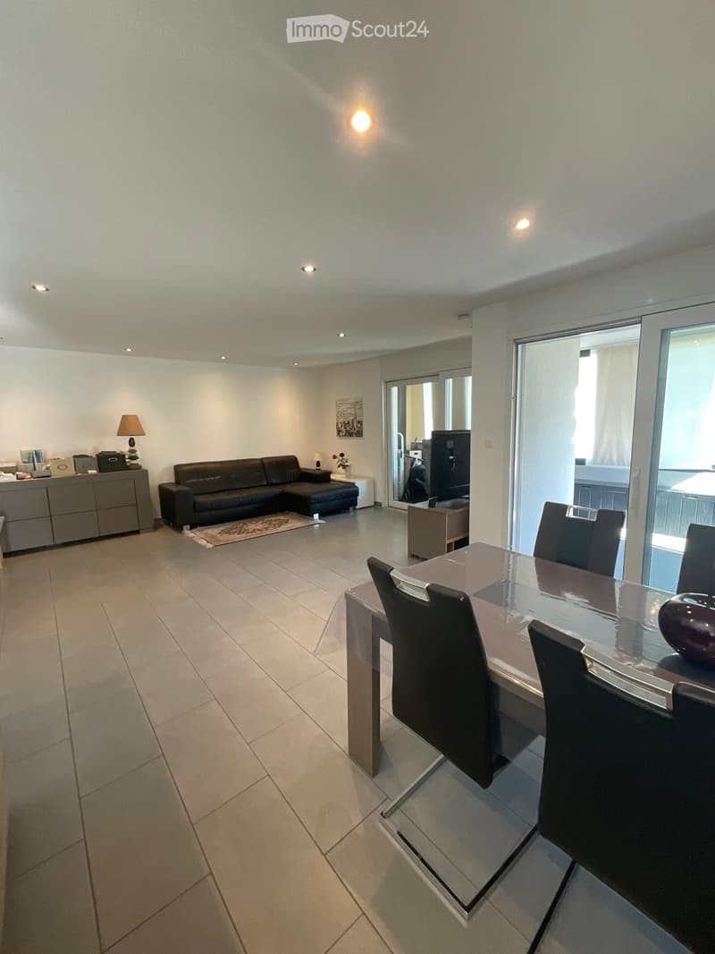 Modern living and dining area with a black leather sofa, glass dining table with chairs, a side cabinet, and a lamp. The space features large windows and a sliding glass door leading to a balcony.