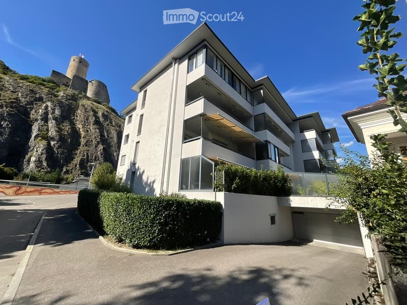 Modern multi-story residential building with white facade, large windows, balconies, and a garage entrance, situated near a rocky hillside with a castle-like structure in the background.