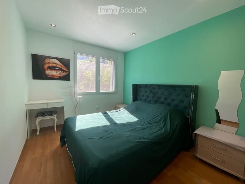 A modern bedroom with a large bed, nightstand, mirror, and a small table with a stool. The room features light blue walls, wooden flooring, and a large window with a view.