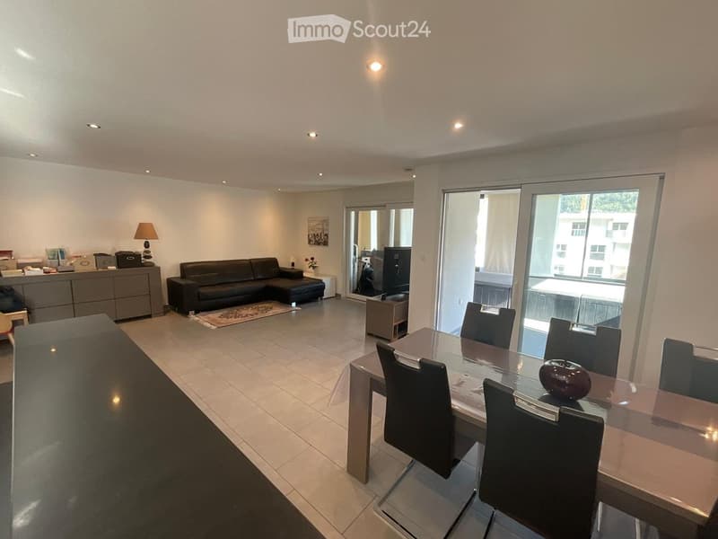 Modern living and dining area with a black leather sofa, glass dining table with chairs, a side table with a lamp, and a cabinet with decorative items.