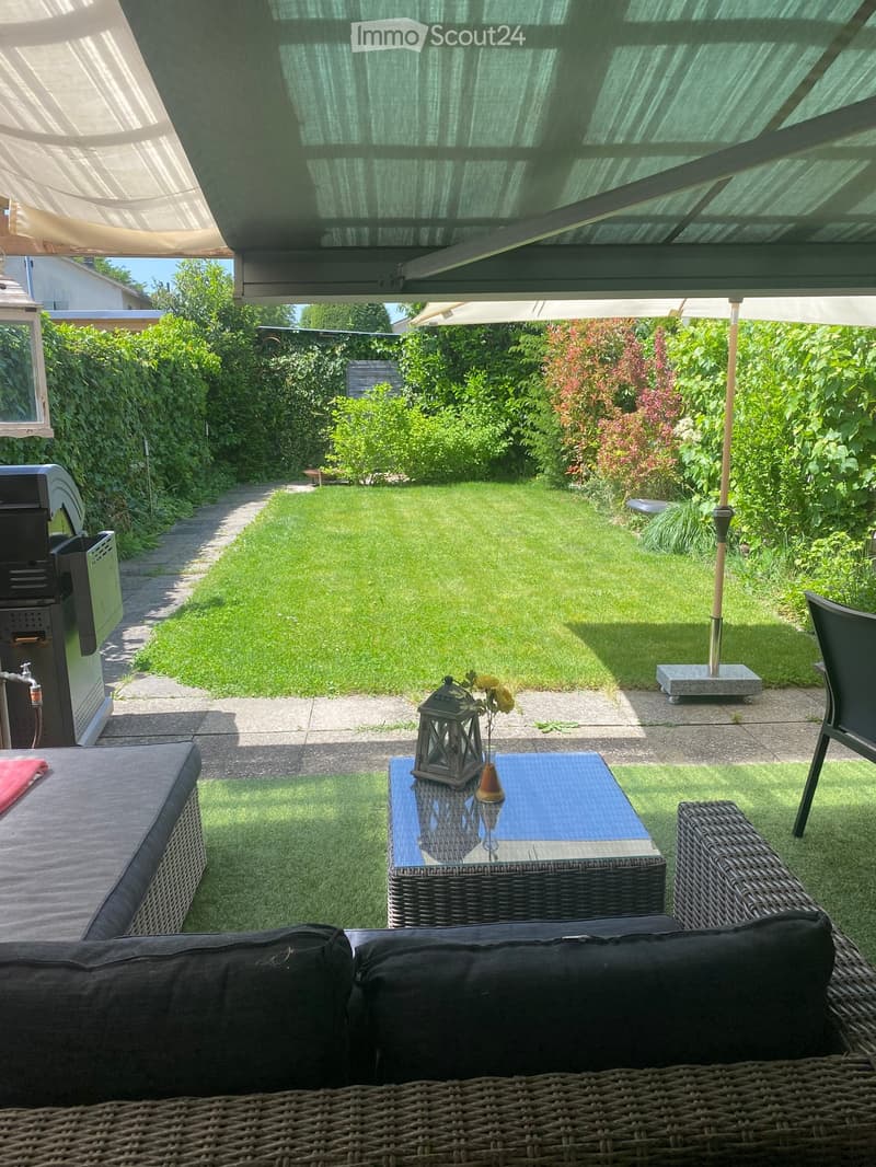 A covered outdoor patio area with wicker furniture, a small table with a lantern and vase, and a well-maintained lawn in the background.