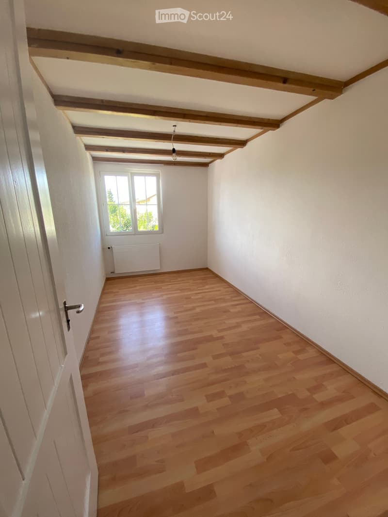 Empty room with wooden flooring, white walls, exposed wooden beams on the ceiling, a single window, and a radiator below it.