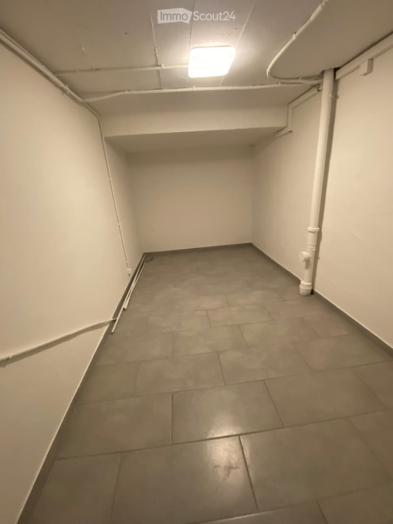 Empty room with tiled floor and white walls, featuring exposed pipes and a ceiling light fixture.