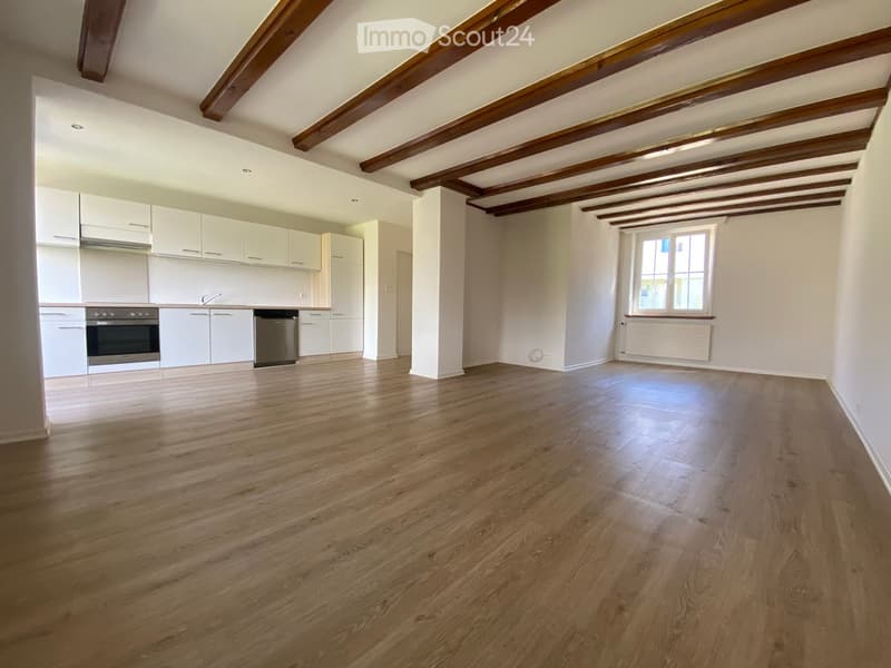 An unfurnished room with wooden flooring, white walls, and exposed wooden beams on the ceiling. A kitchen area with white cabinets and appliances is visible in the background, along with a window and radiator on one wall.