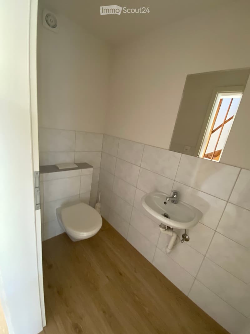 Small bathroom with white tiled walls, wooden floor, toilet, sink, and a small window above the sink.