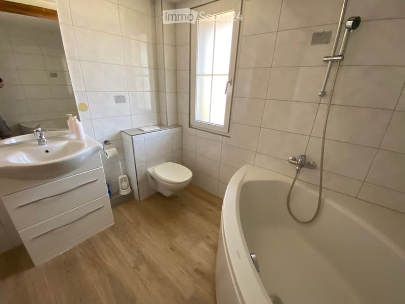 Modern bathroom with white tiles, wooden flooring, a bathtub with a handheld shower, a toilet, a sink with a mirror, and a window for natural light.