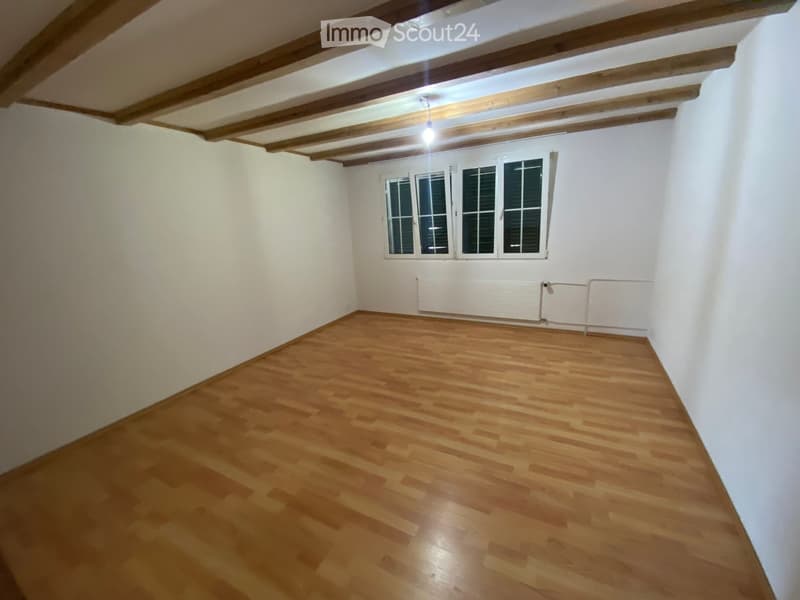 Empty room with wooden flooring, white walls, and exposed wooden ceiling beams. Features a window with shutters and a radiator below it.
