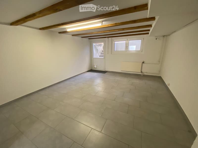 Empty room with tiled floor, white walls, exposed wooden beams on the ceiling, a door, and a window with two panes.