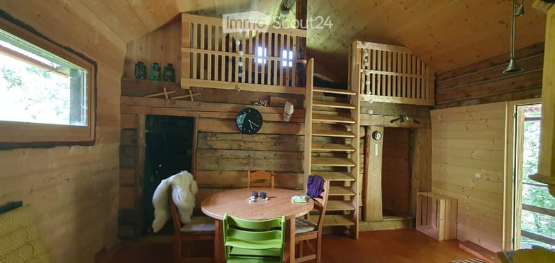 Cozy wooden cabin interior with a round dining table, chairs, a futon on an elevated platform, shelves, and decorative vases.