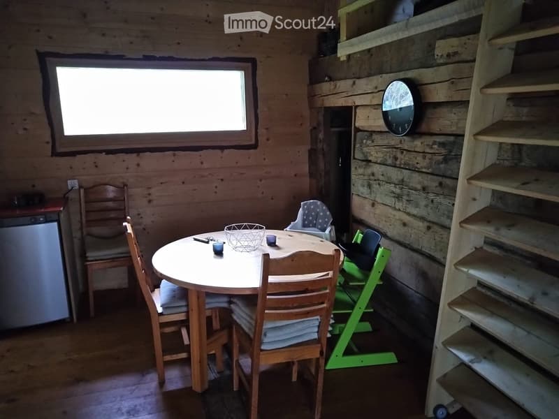 A cozy wooden cabin interior with a round wooden dining table surrounded by chairs, a baby chair, a refrigerator, and a clock on the wall.
