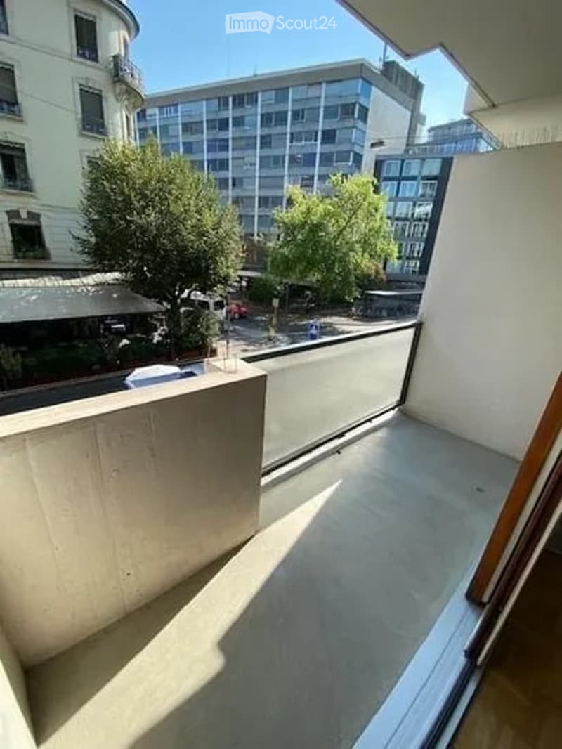 Balcony with a modern design, featuring a concrete floor and glass railing, overlooking a cityscape with trees and buildings.