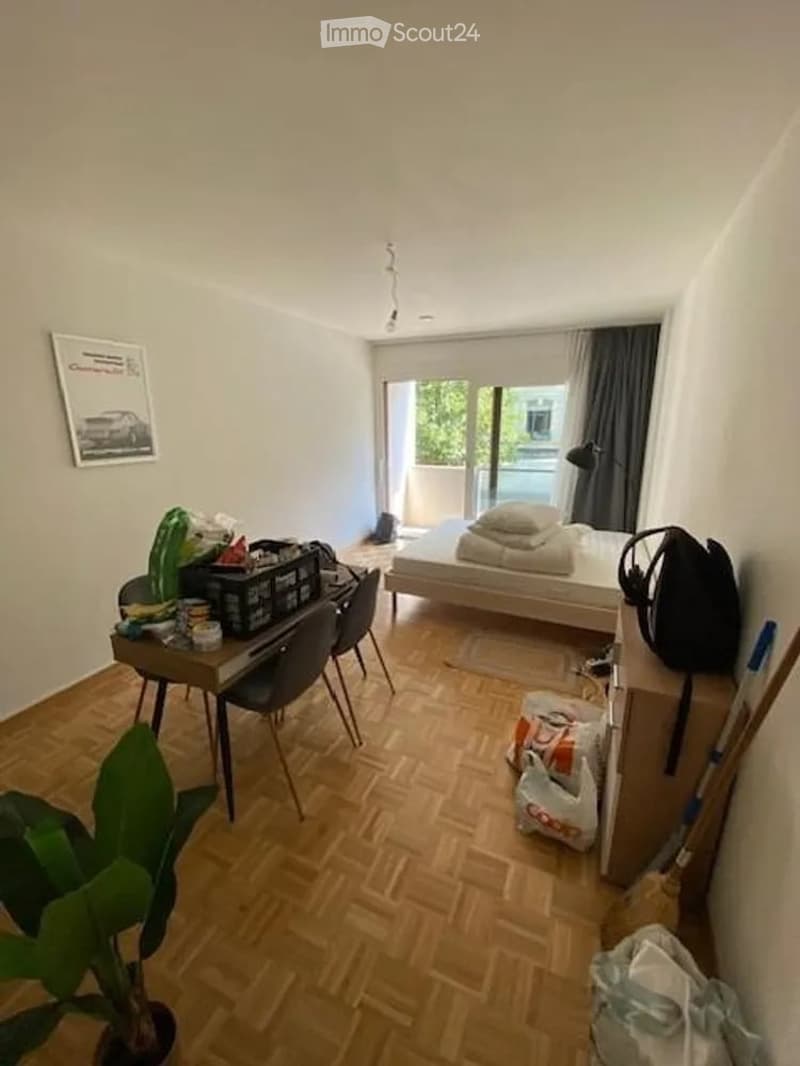 Cozy bedroom with wooden flooring, a bed with pillows and blankets, a small table with chairs, a lamp, and a balcony with a view of greenery.
