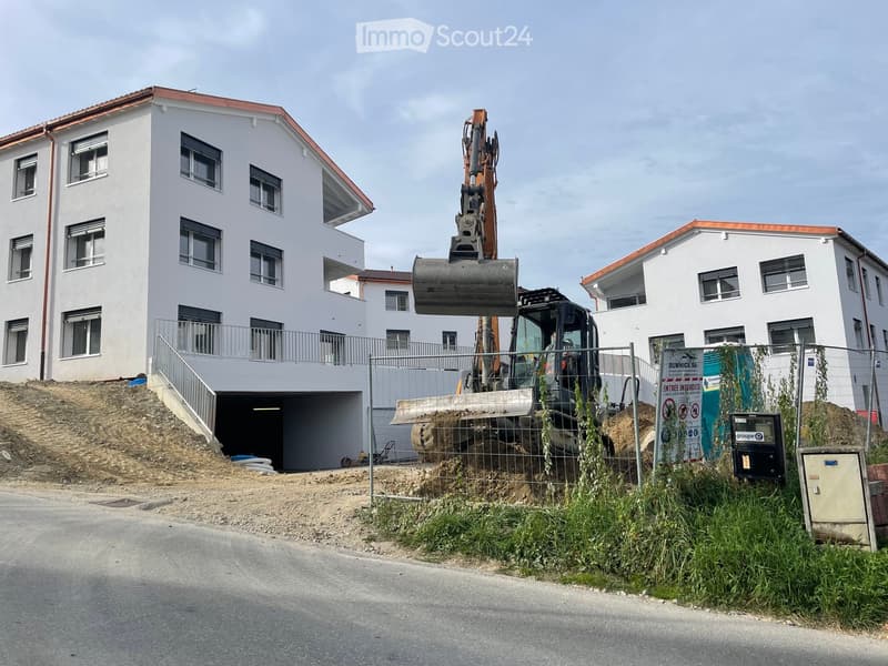 Two-story white residential building with a red roof, multiple windows, and a fenced-off construction area with an excavator in the foreground.