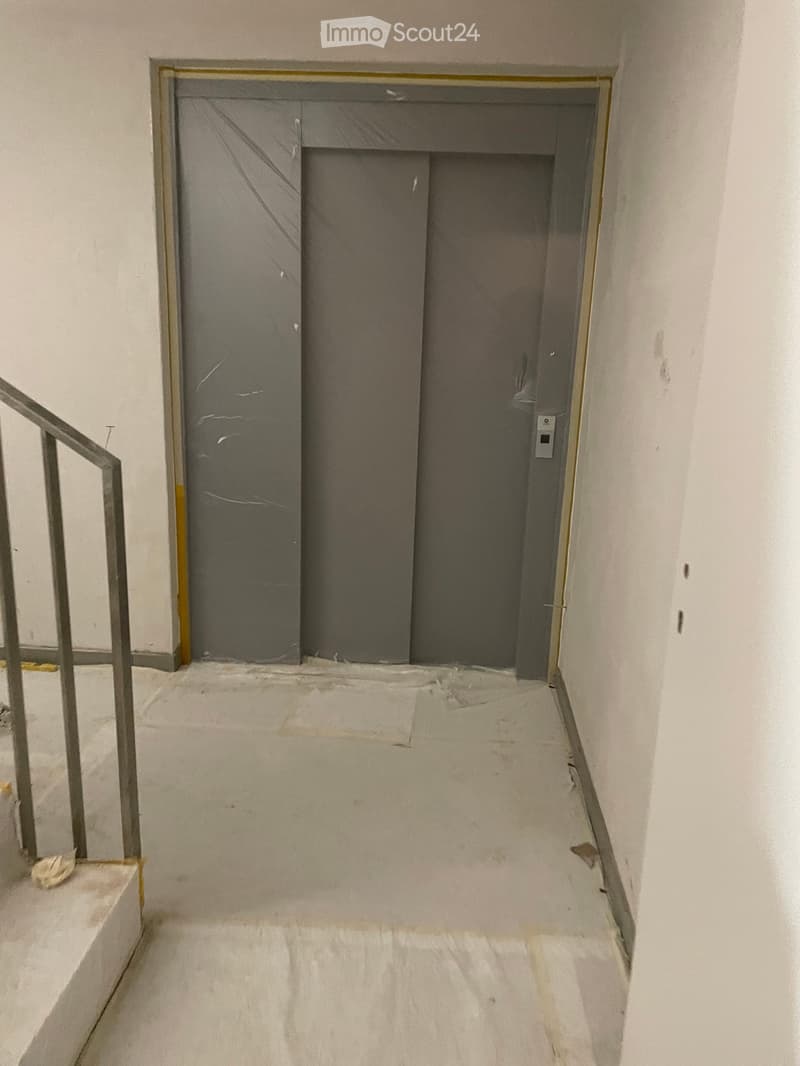 Empty hallway with white walls, a metal staircase railing on the left, and a closed elevator door at the end.