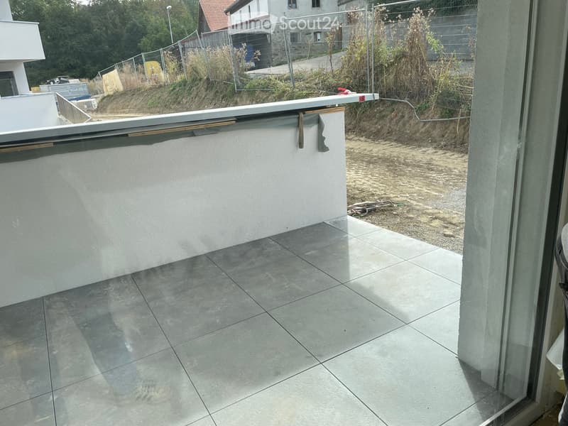 Balcony with tiled floor, white railing, and view of a fenced area with construction materials and greenery.