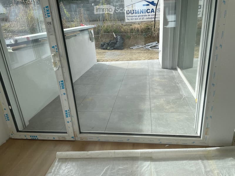 Empty room with wooden flooring and large glass doors leading to a tiled patio area. The room is unfurnished and appears to be under construction or renovation, with protective plastic sheeting on the floor and construction materials visible outside.