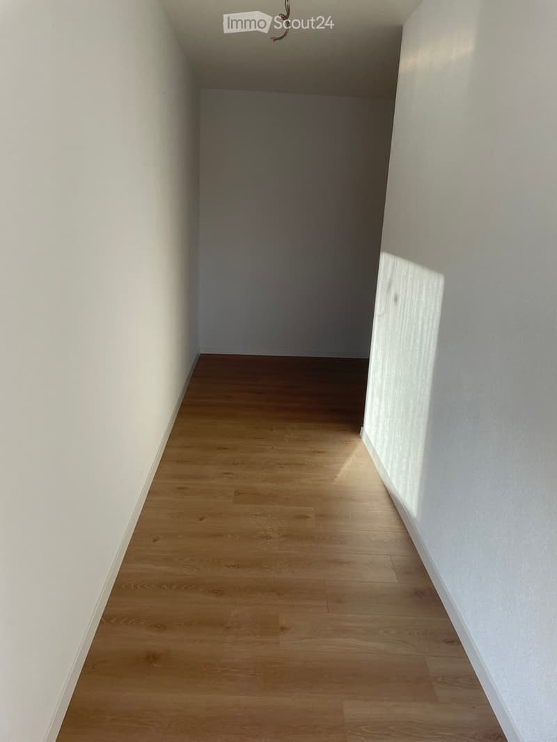 Empty hallway with light wooden flooring and white walls, leading to an open doorway.
