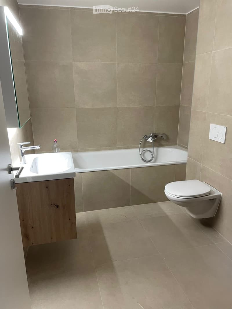 Modern bathroom with beige tiles, a white sink with a chrome faucet, a bathtub with a handheld shower, a toilet, and a mirror on the wall.