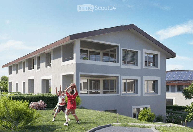 Modern two-story house with light gray exterior, large windows, and a balcony on the upper floor. The house features a sloped roof with dark roofing material and is surrounded by a well-maintained lawn and garden area.