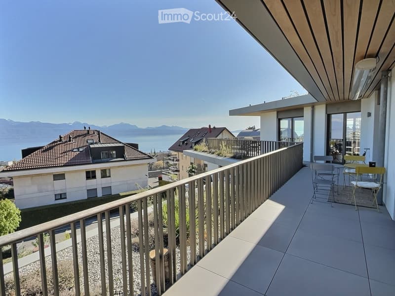 Balcony with tiled floor, metal railing, and outdoor furniture including chairs and a table, overlooking a scenic lake and mountain view.
