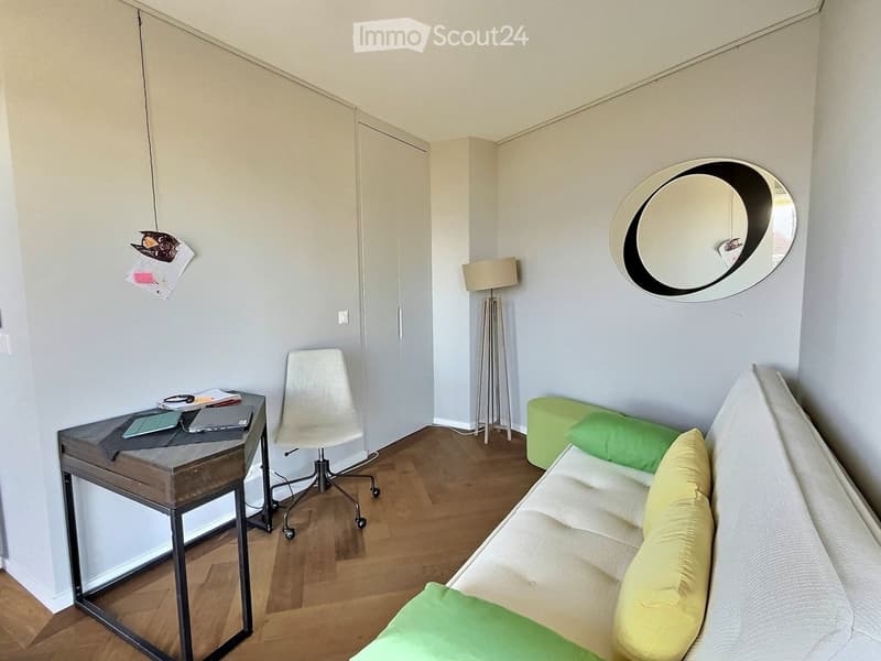 A modern, well-lit room with a sofa, a chair, a small table with a laptop, a floor lamp, a round mirror, and a green ottoman.