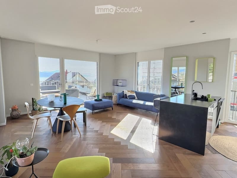 Modern living room with wooden flooring, large windows offering a scenic view, a blue sofa, a coffee table, and a kitchen island with a sink.