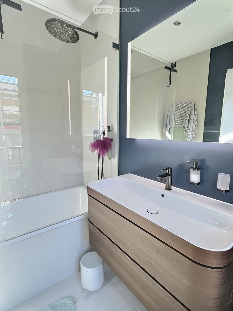 Modern bathroom with a white sink, wooden vanity, large mirror, bathtub, and showerhead. Includes a toilet, soap dispenser, and towel.