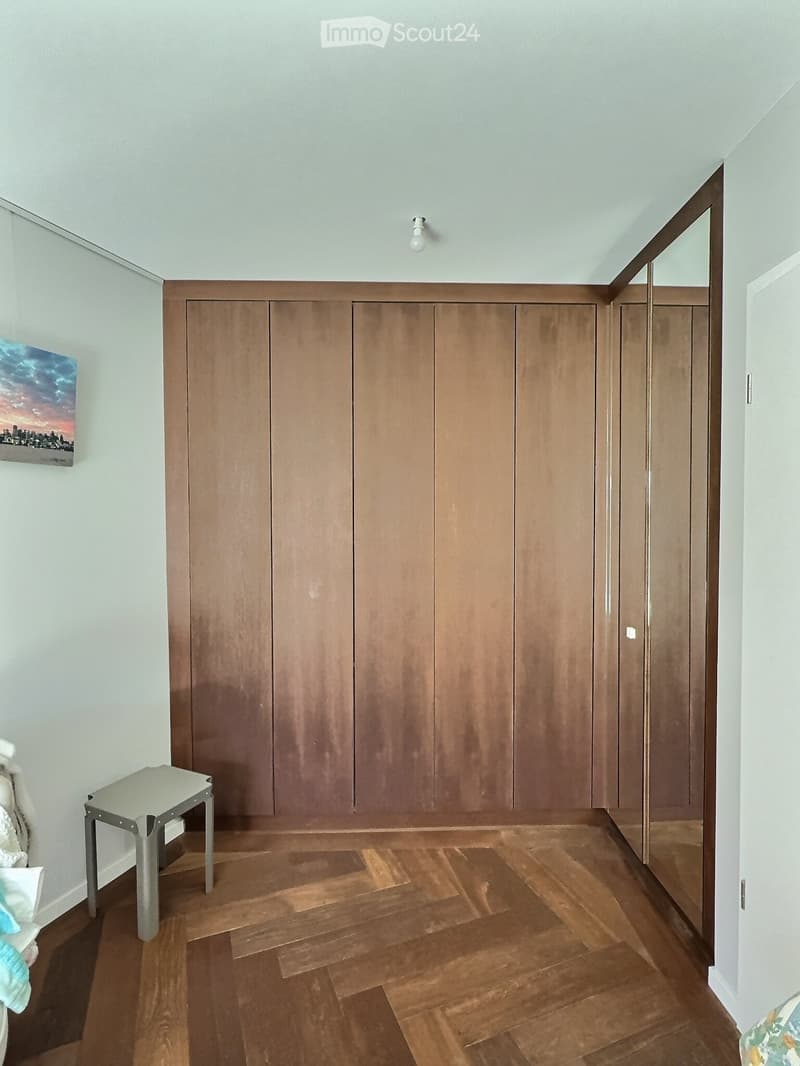 A small, furnished room with wooden flooring, built-in wooden wardrobes, a small grey stool, a mirror, and a painting on the wall.