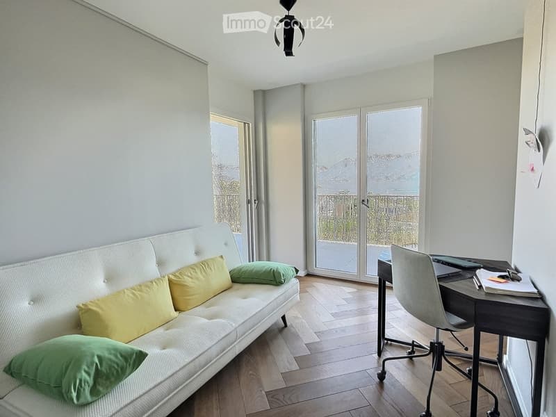 A modern, well-lit room with a white sofa, green cushions, a desk with a chair, and a laptop. Large windows and sliding glass doors lead to a balcony.