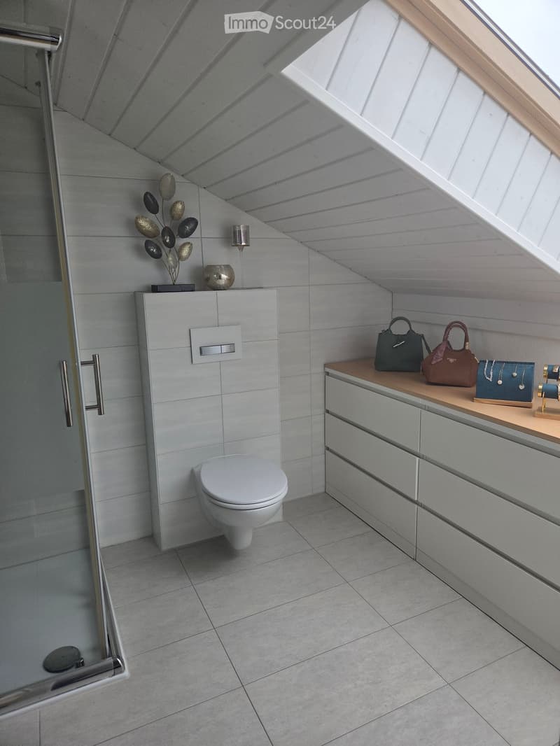 Modern bathroom with a toilet, mirrored cabinet, wooden countertop, decorative items, and handbags on display.