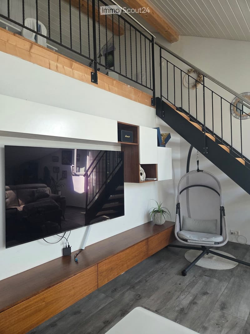 Modern living room with a large flat-screen TV mounted on the wall, a wooden entertainment unit, a grey rocking chair, and a small plant on the table. The room features a staircase leading to an upper level.