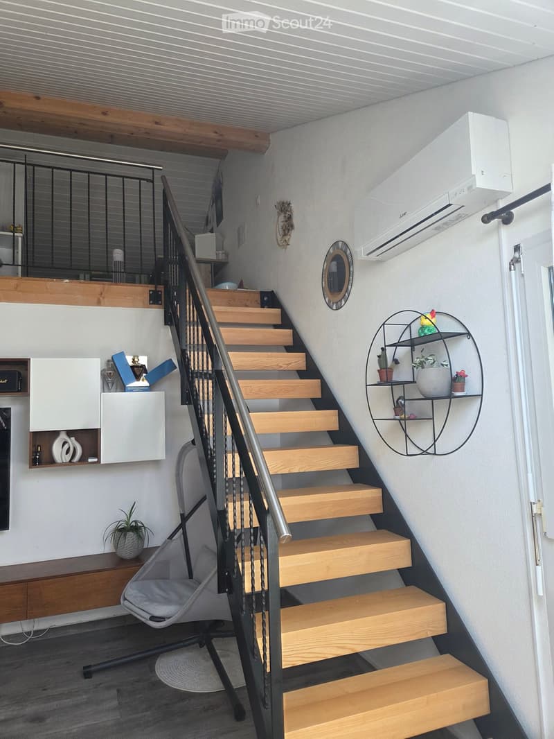 Staircase with wooden steps and metal railing, small seating area with a chair, shelves with decorative items, and a plant on a table.
