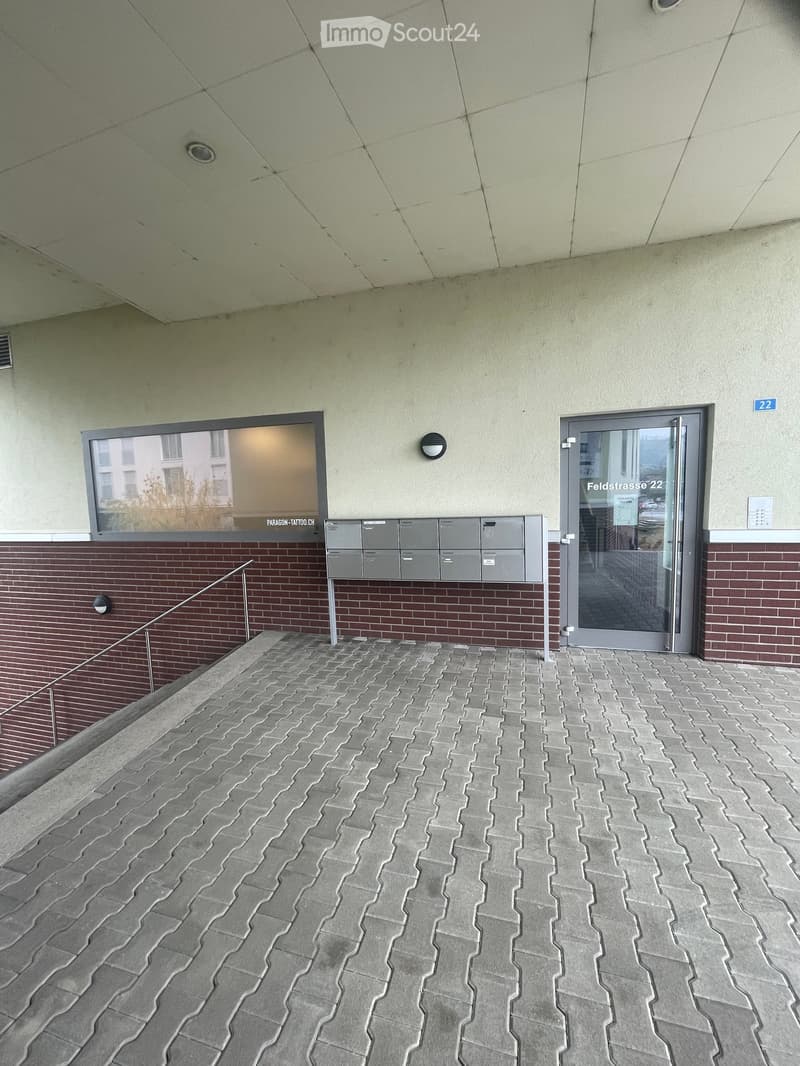 Balcony with gray brick pavement, modern glass door labeled "Feldstrasse 22", mailboxes, and a window showing a building view.