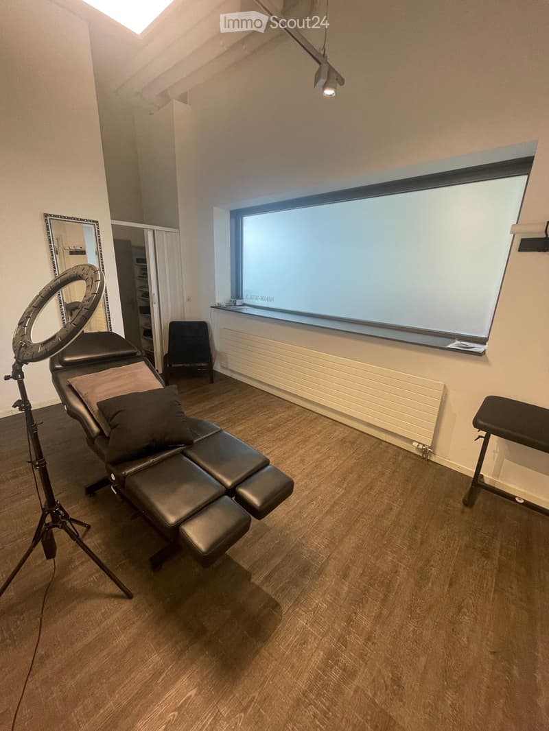 A well-lit room with wooden flooring, a large window, a black massage chair with pillows, a small table, a mirror, and a stool.