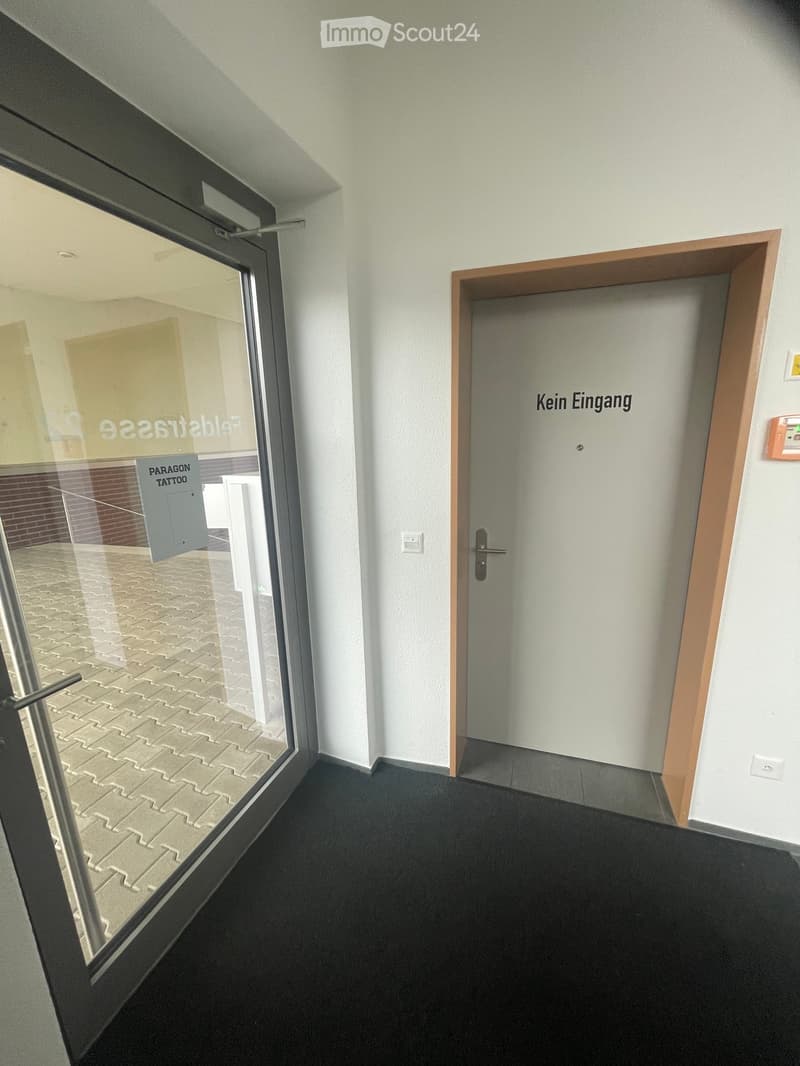Empty hallway with a glass door on the left and a closed door labeled "Kein Eingang" on the right. The floor is dark carpeted, and the walls are white.