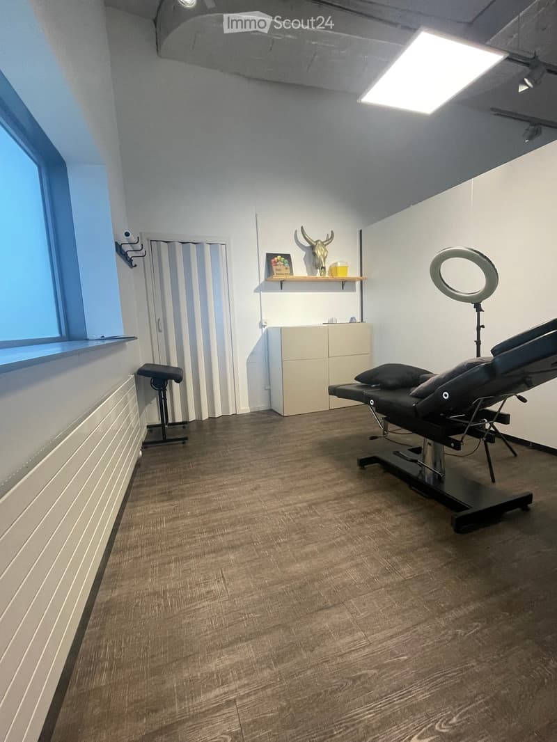 Modern, well-lit room with wooden flooring, a black massage table, a round mirror on a stand, a small shelf with decorative items, and a white cabinet.
