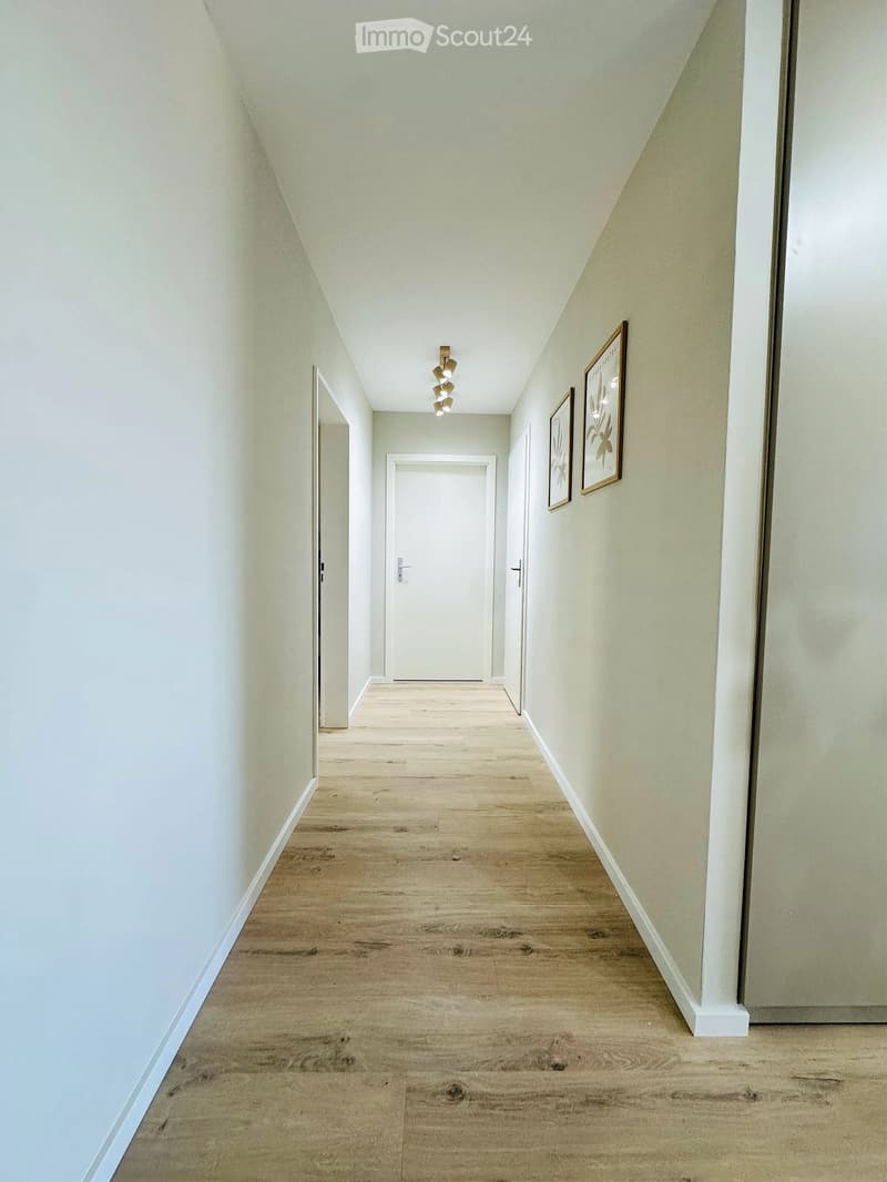 A narrow hallway with light-colored wooden flooring, white walls, and a ceiling light fixture. Two framed pictures are mounted on the right wall, and there are three doors along the hallway.