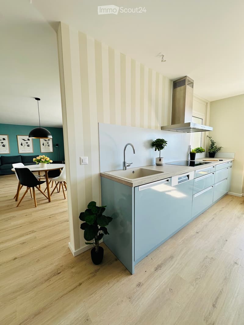 Modern kitchen with light blue cabinets, a central island, a sink, a dishwasher, and a range hood. The space features wooden flooring, pendant lighting, and a small dining area with a table and chairs visible in the background.