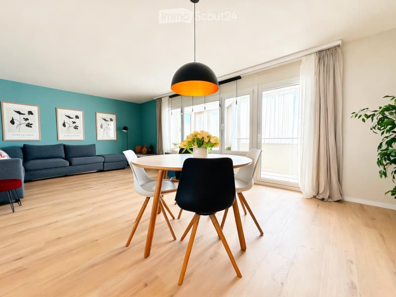 Modern living and dining area with light wood flooring, teal accent wall, white dining table with four chairs, grey sofa, pendant light, and large windows with sheer curtains.
