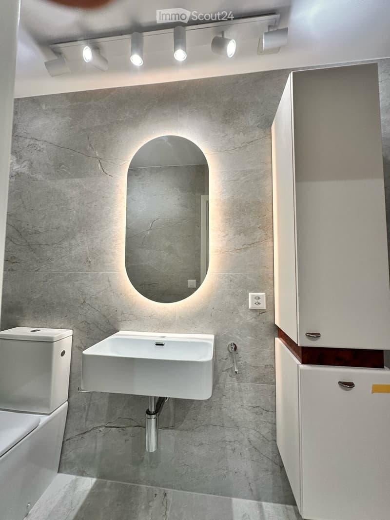 Modern bathroom with a white sink, toilet, and built-in cabinet. Features a large, oval mirror with integrated lighting and sleek, grey marble tiles.
