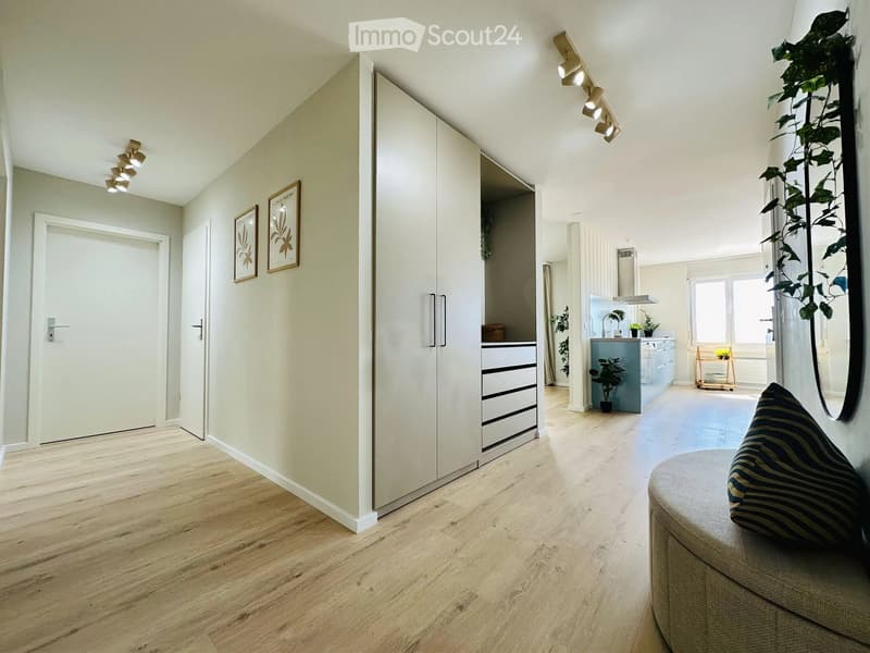 Modern, open-plan living and dining area with light wood flooring, white walls, and contemporary furniture including a sofa, potted plants, and a kitchen area with cabinets and a sink.