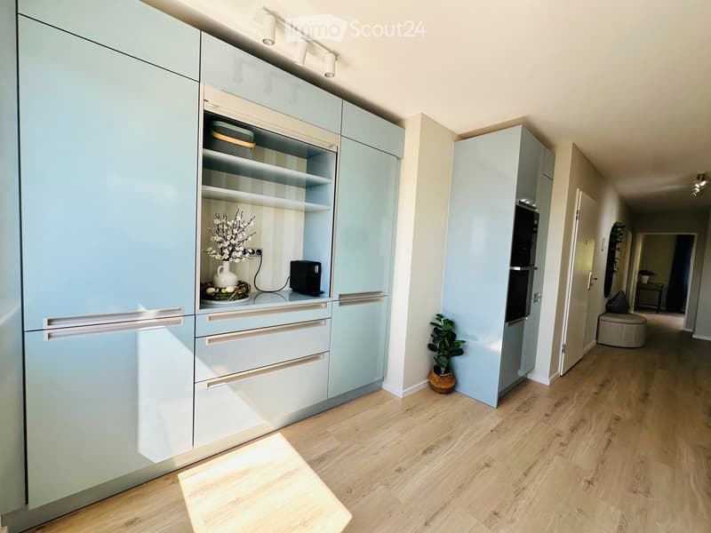 Modern kitchen with light blue cabinets, stainless steel handles, and a wooden floor. Includes a refrigerator, oven, and a small table with a vase and plant.