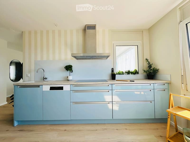 Modern kitchen with light blue cabinets, a stainless steel sink, a dishwasher, and a range hood. The countertops are light-colored, and there are small potted plants on the windowsill and a wooden stool near the window.