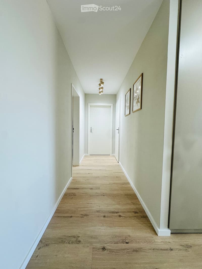 A clean, modern hallway with light wooden flooring, white walls, and a ceiling light fixture. Two framed pictures are mounted on the right wall, and there are three doors along the corridor.